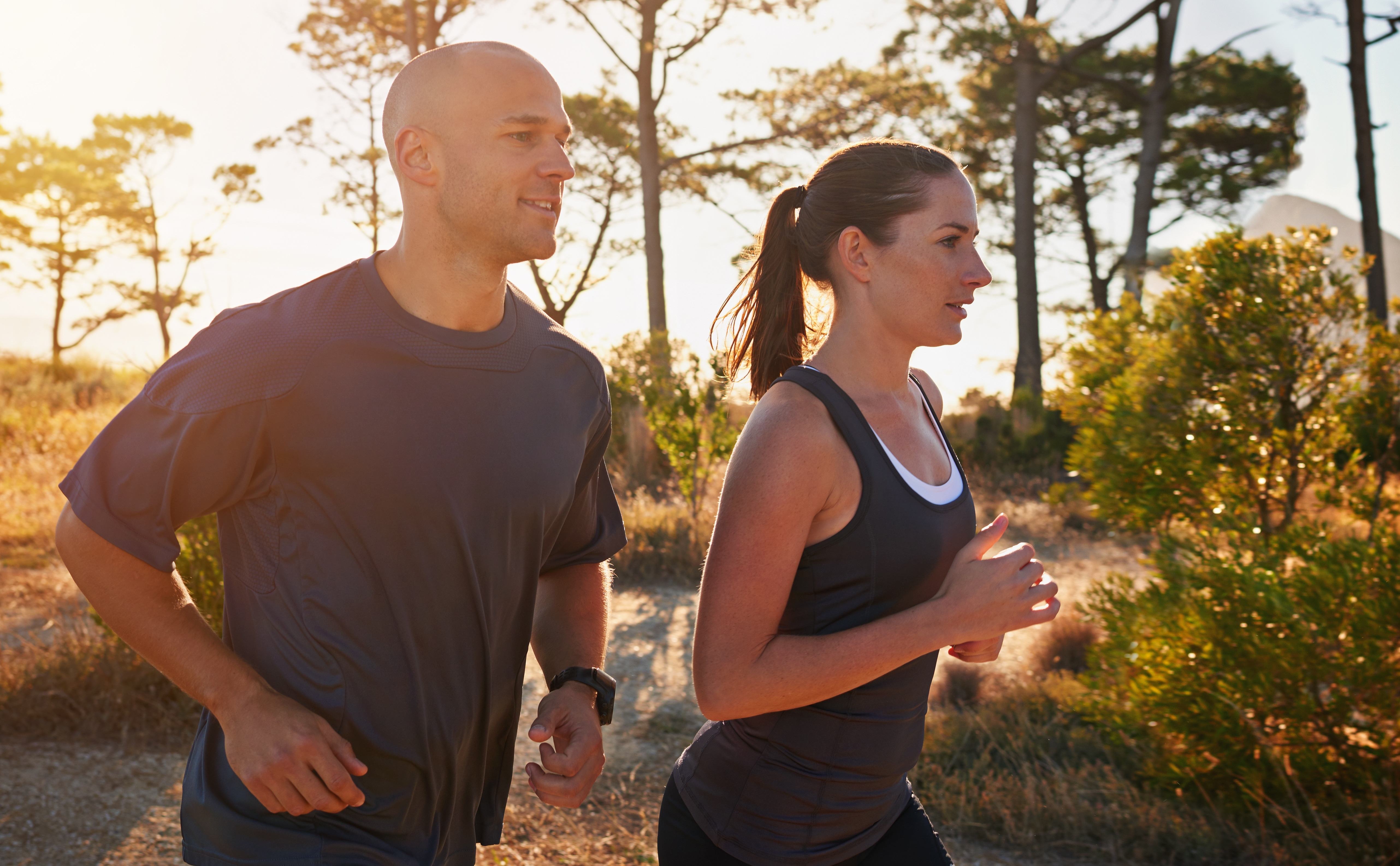 A man and woman jogging together on a sunlit outdoor trail surrounded by trees and greenery.